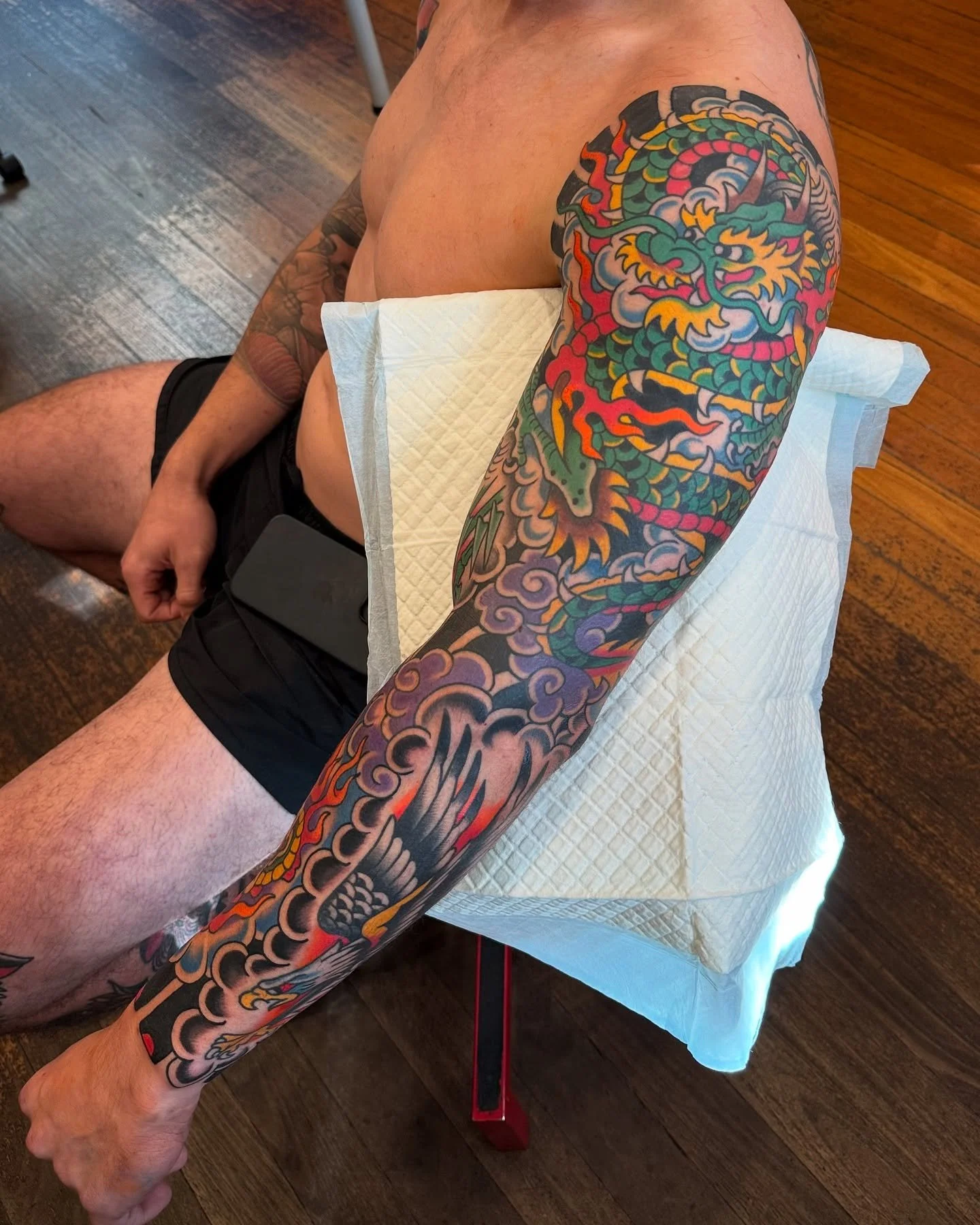 A man with colorful Japanese-style dragon tattoos on his arm, sitting on a tattoo chair with a paper towel draped over his lap in a tattoo studio.