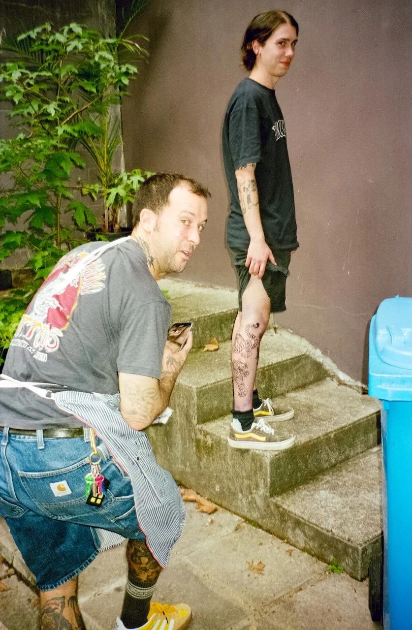 Two men with tattoos, one crouching and holding a phone, the other standing on outdoor steps, near a green plant, a brown wall, and a blue recycling bin.