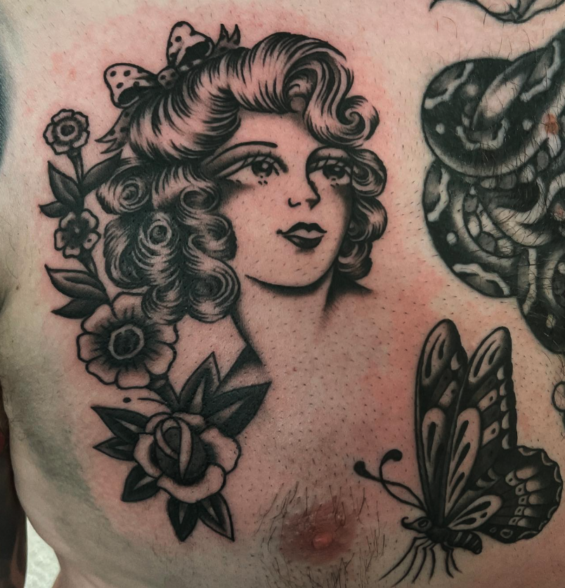 Tattoo of a woman's face with curly hair, surrounded by flowers, a butterfly, and a snake, on a person's chest.