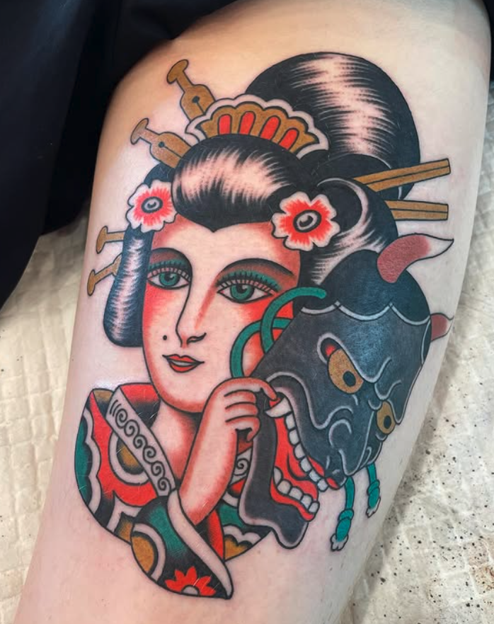 Tattoo of a traditional-style woman with black hair and flowers, holding a stylized blue mask with fierce facial features, surrounded by ornamental details.