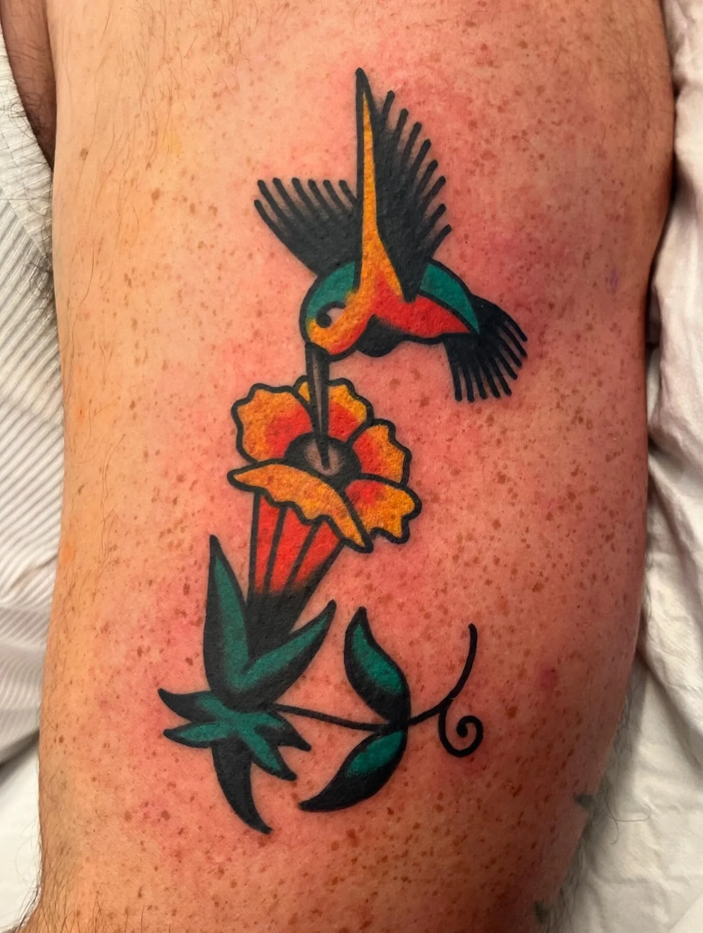 Colorful tattoo of a hummingbird drinking nectar from a flower, with green leaves and a black outline.