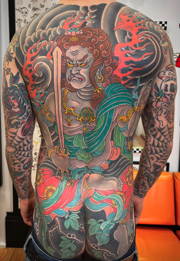 A person with extensive traditional-style tattoos on their back and arms, featuring a fierce warrior or deity with a lion's face, holding a sword, with flowing robes, clouds, and decorative elements in vibrant colors.