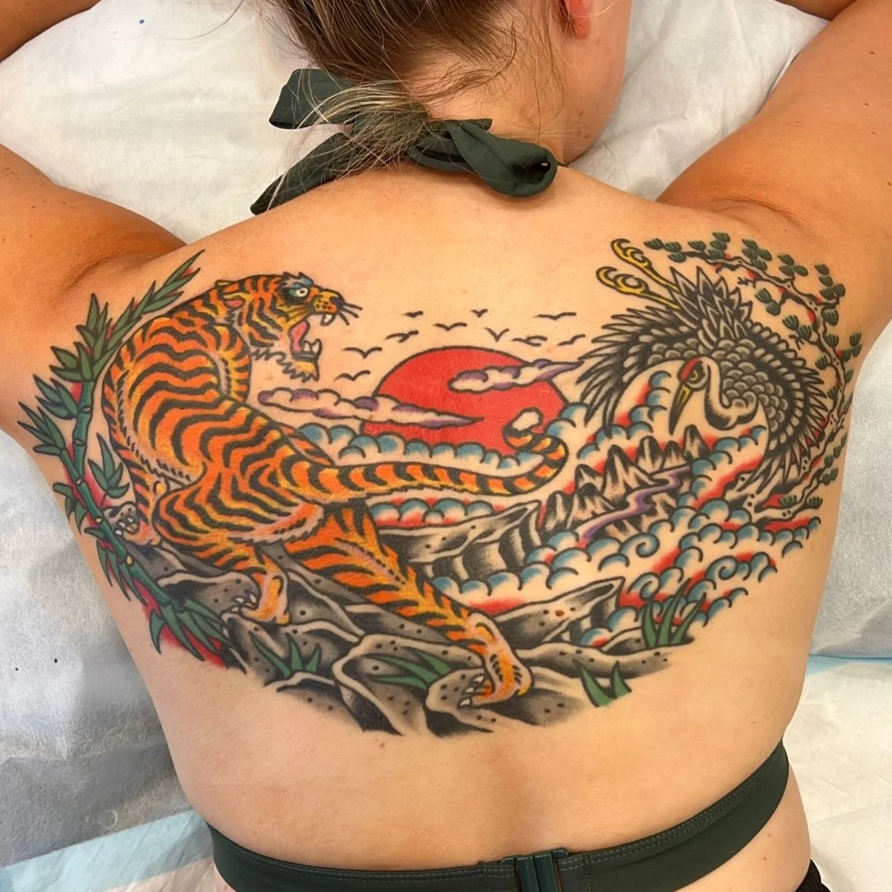 Colorful tattoo of a tiger and a phoenix on a person's back with a red sun, clouds, and birds in the background.