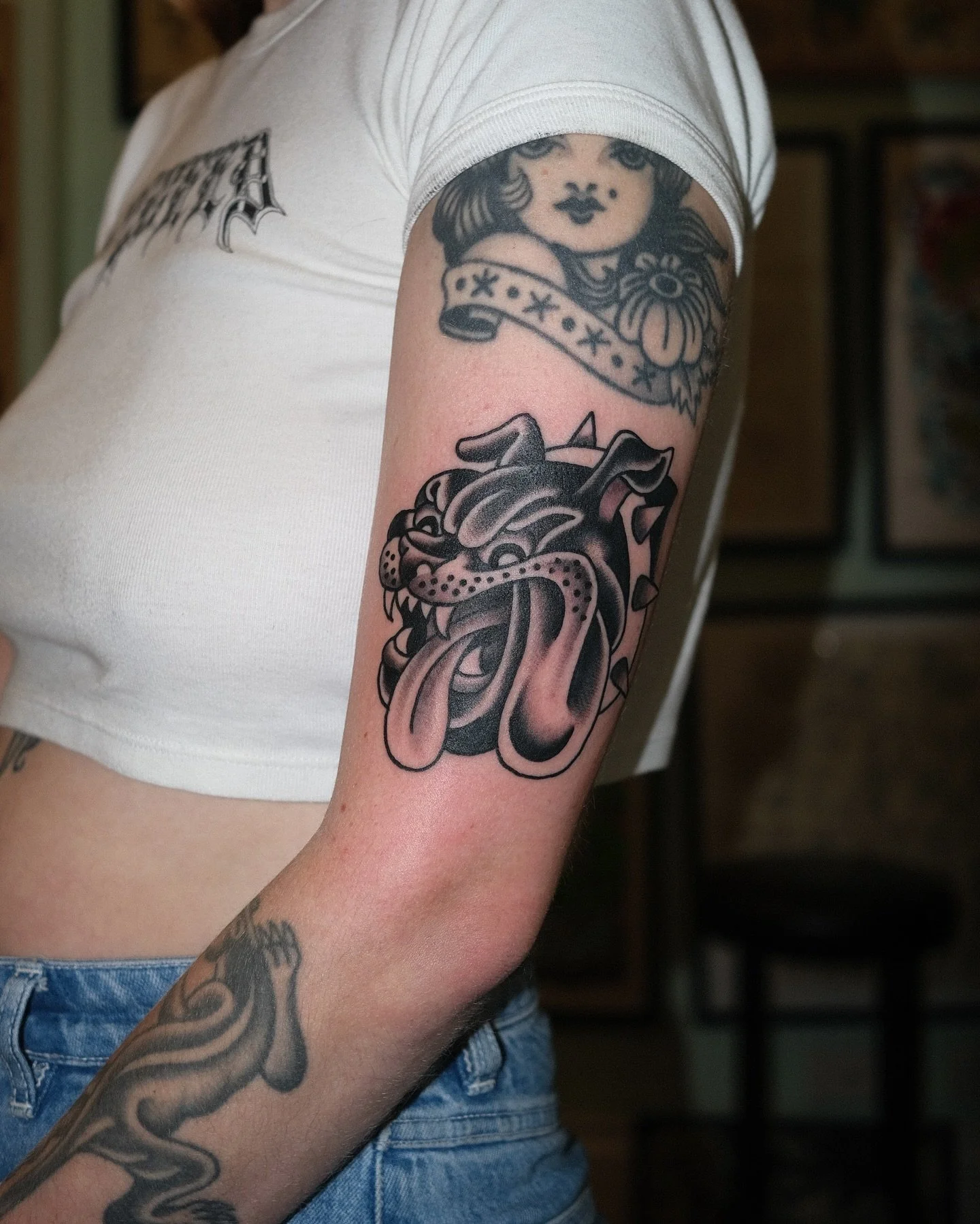 Tattoo of an aggressive, snarling dog with an open mouth and protruding tongue on person's upper arm, with a tattoo of a woman with dark hair and banner above on upper arm.