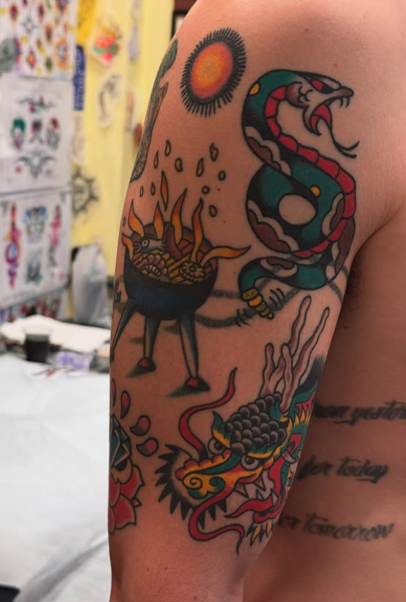 Tattoo of a snake, a pot with flames, a sun, and a dragon on person's arm.
