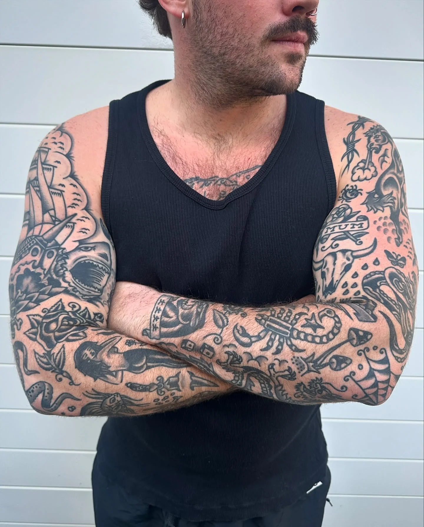 A man with tattoos on both arms, wearing a black sleeveless shirt, standing with arms crossed against a light-colored wall.