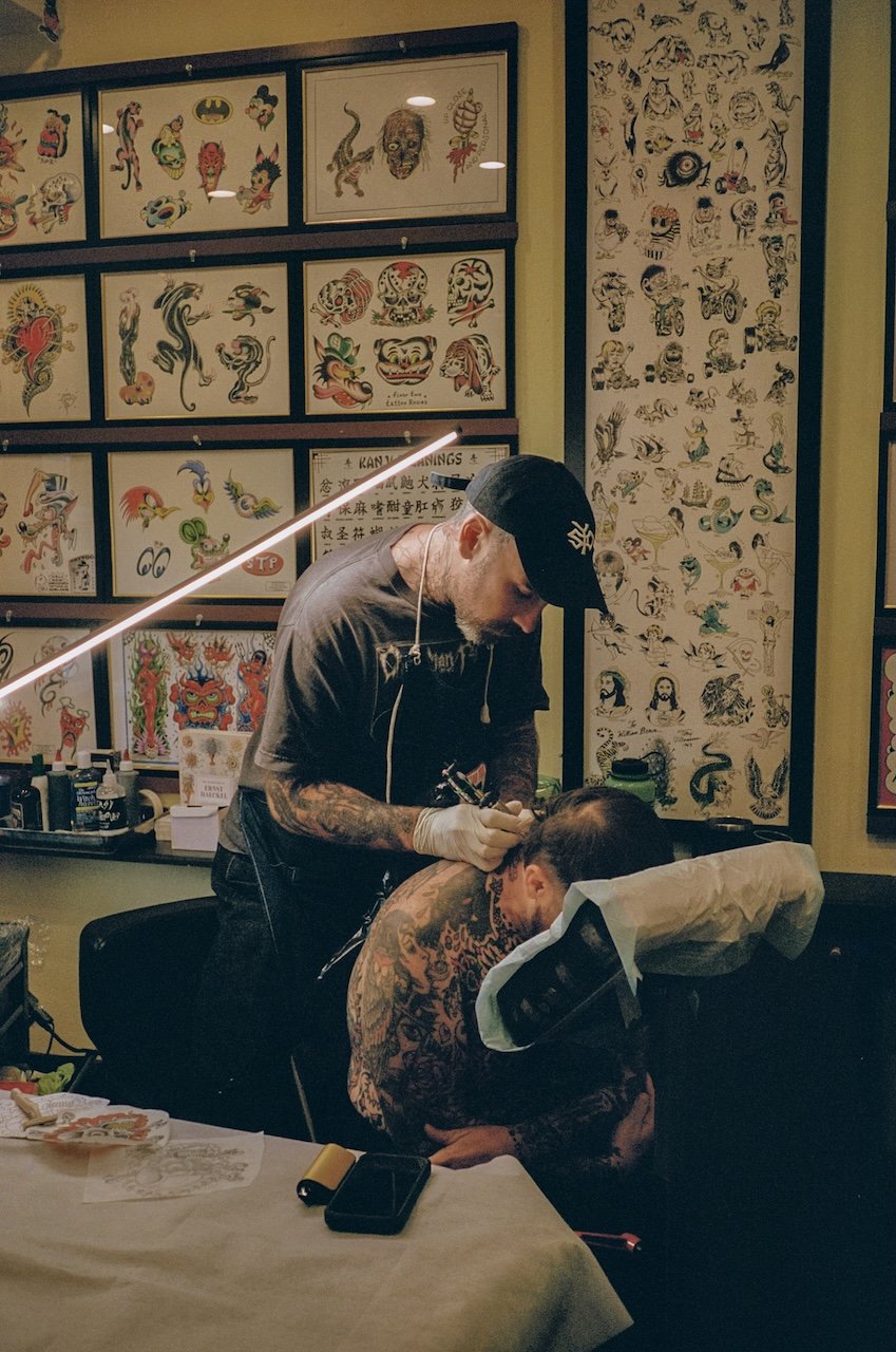 A tattoo artist with tattoos on his arms tattooing a man's back in a tattoo studio, decorated with tattoo flash art on the walls.