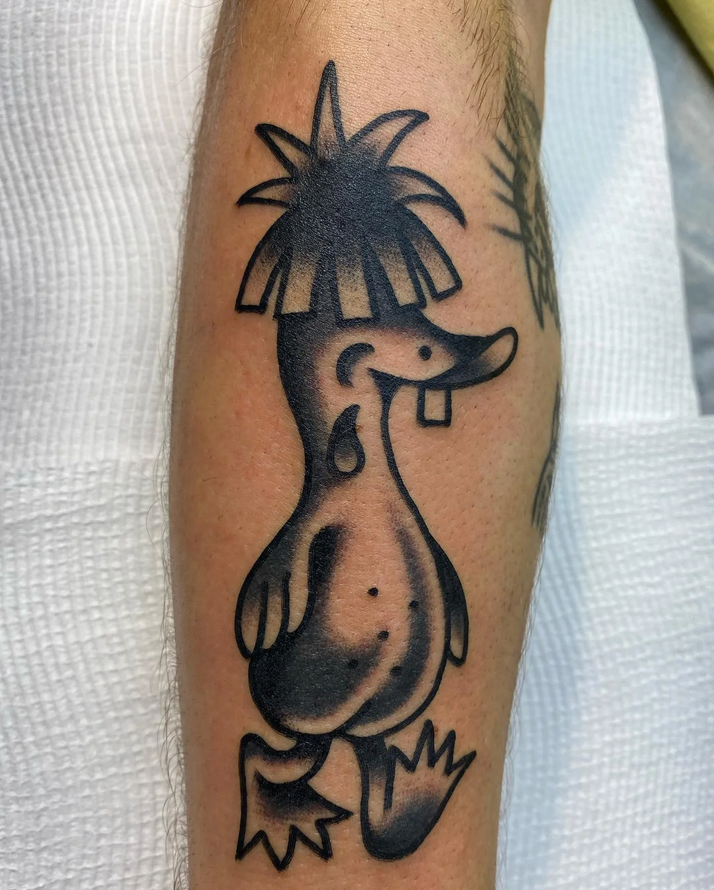 A black tattoo of a cartoon-style narwhal with a big smile, wearing a pineapple on its head, with a small splash near its tail.