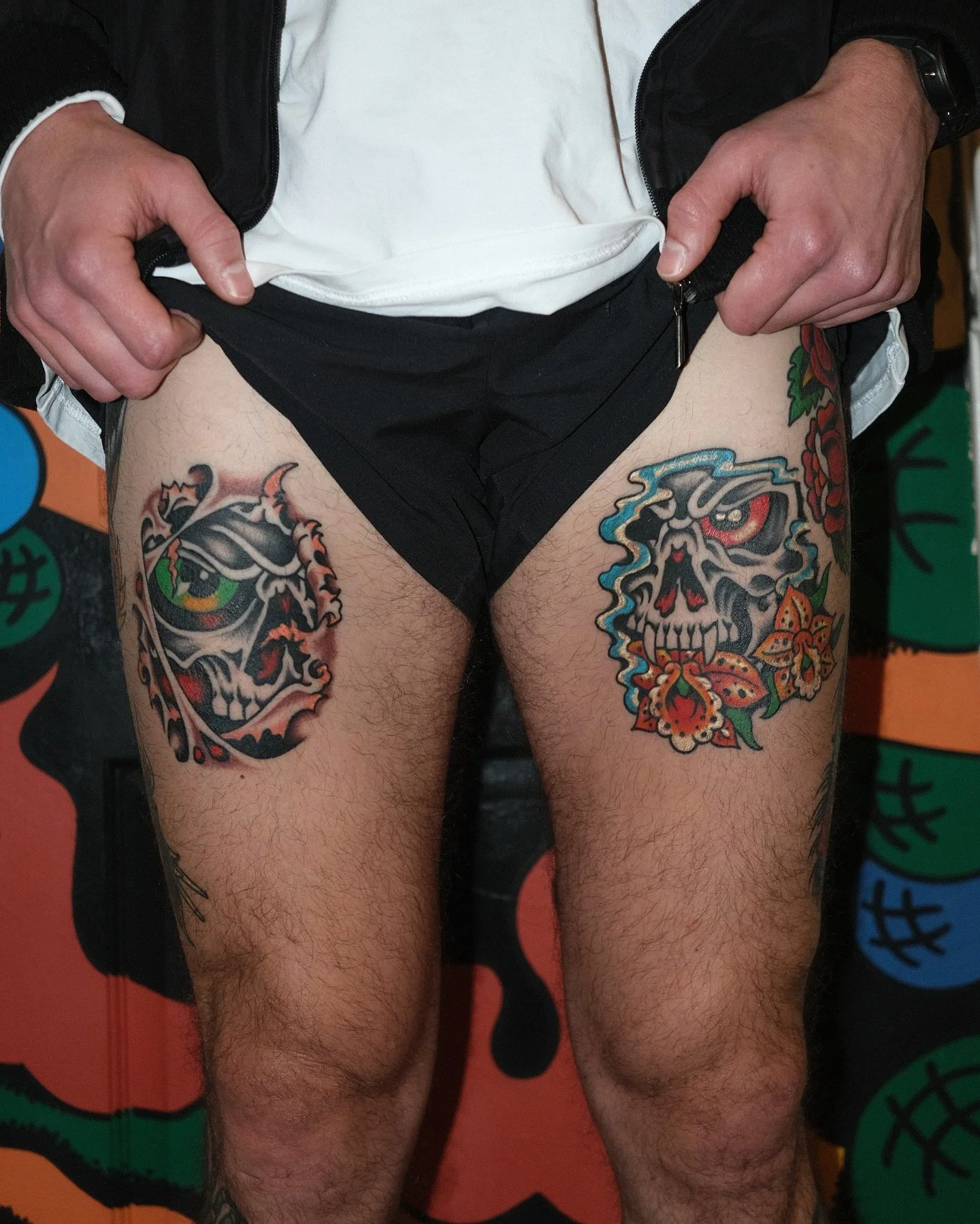 A person with tattoos on their thighs, wearing black shorts and a white shirt, holding open a black jacket to reveal the tattoos.