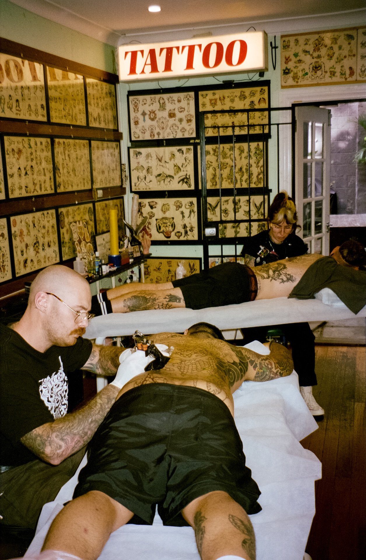 Tattoo artists working on clients in a tattoo shop, with tattoo designs displayed on the walls and a "TATTOO" sign illuminated above.