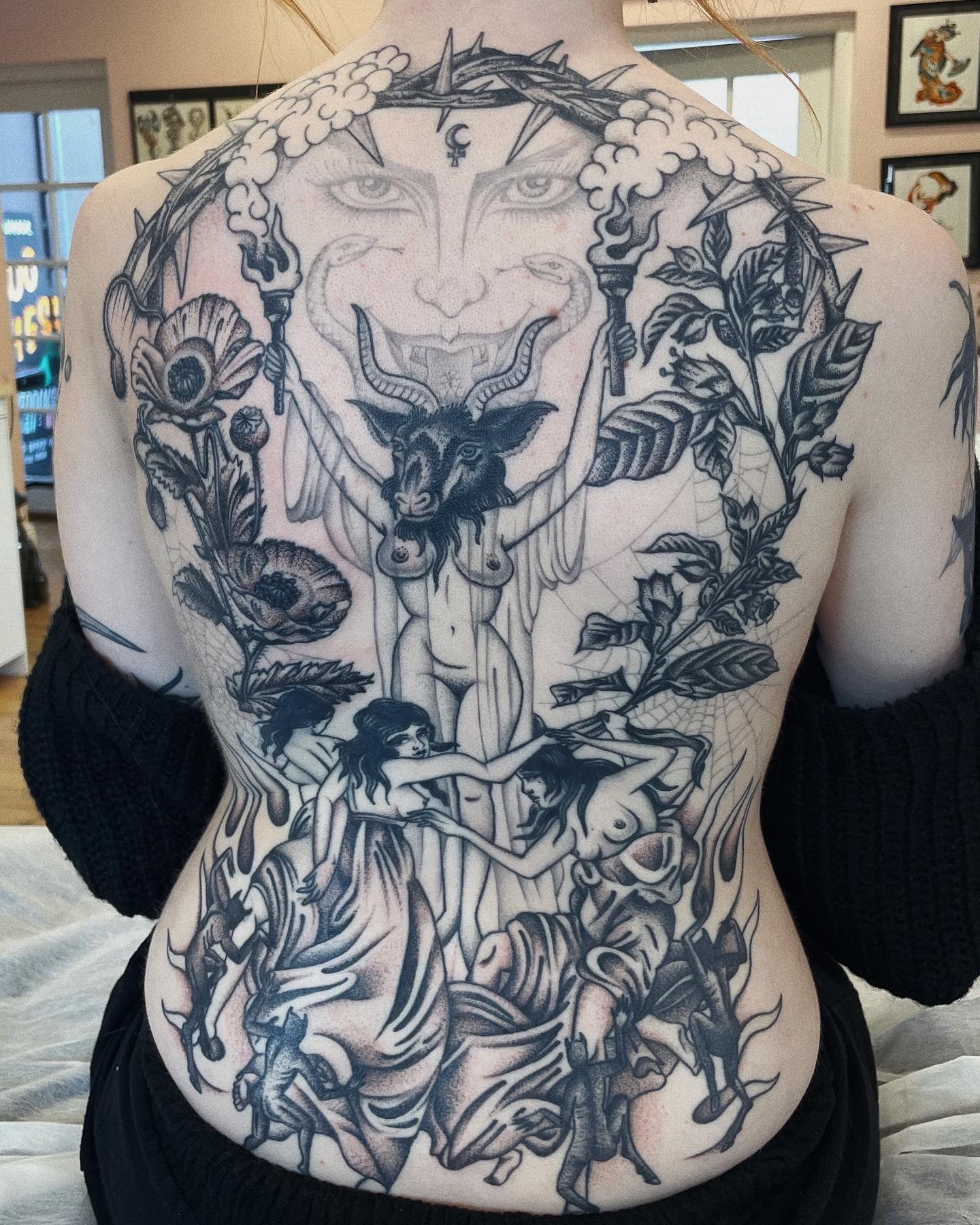 Black and gray tattoo on a person's chest and abdomen, depicting a mythological scene with a goat-headed figure, women, plants, and a smiling face with cloud, crown of thorns, and torches.