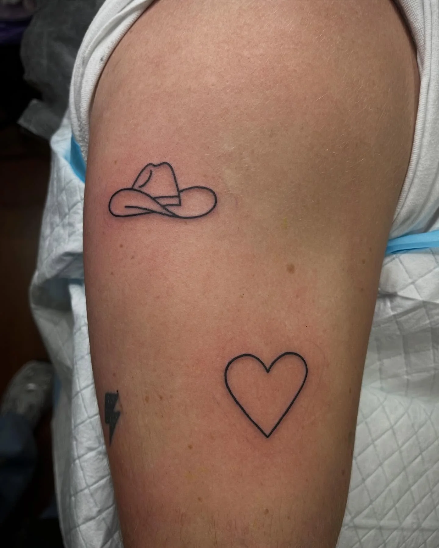 A person's upper arm with two minimalist tattoos: a black line drawing of a cowboy hat and a black outline of a heart.