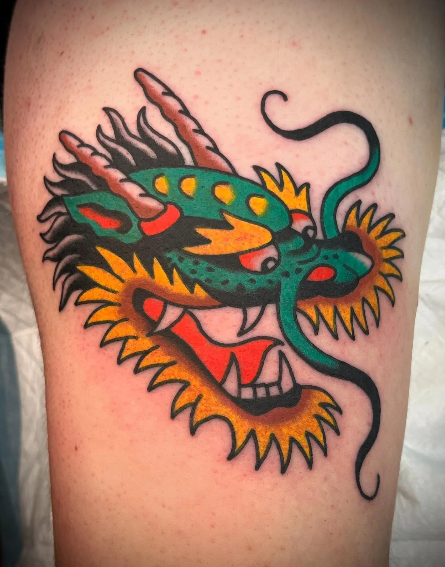 Colorful tattoo of a fierce dragon with a skull at the base of its neck, featuring orange, green, red, and black ink on skin.