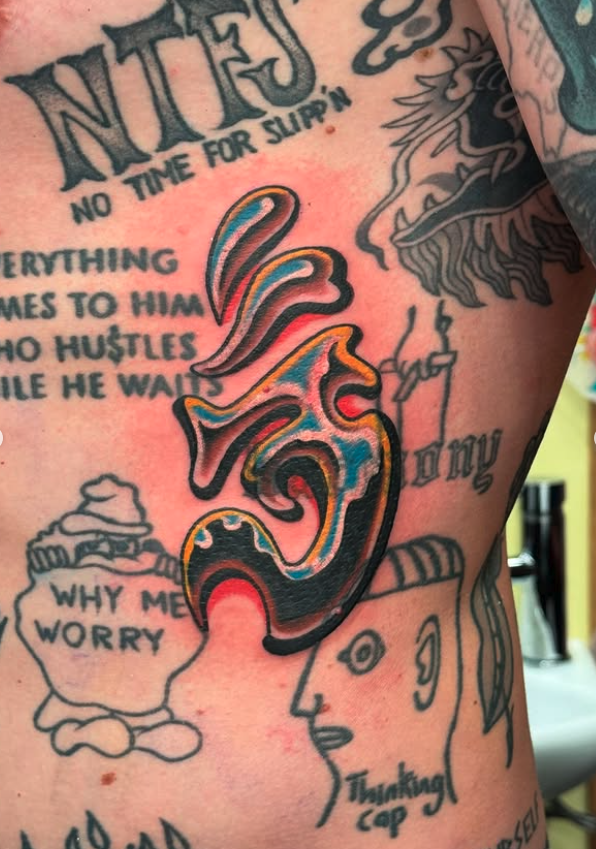 Close-up of a colorful tattoo on a person's chest featuring a stylized face with a substance swirling out of its nose, alongside various black tattoos including a cupcake with the text "Why me worry," a face mask, and text with phrases like "No time 