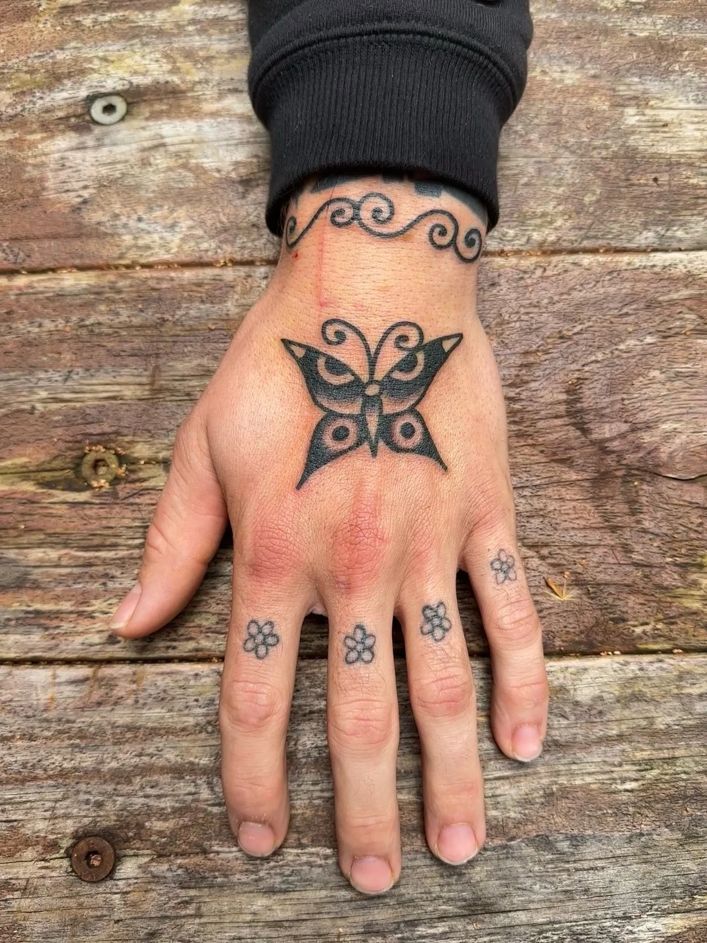 Hand with several tattoos, including a large butterfly on the hand, cloud-like design around the wrist, and small flower tattoos on the fingers, resting on a wooden surface.