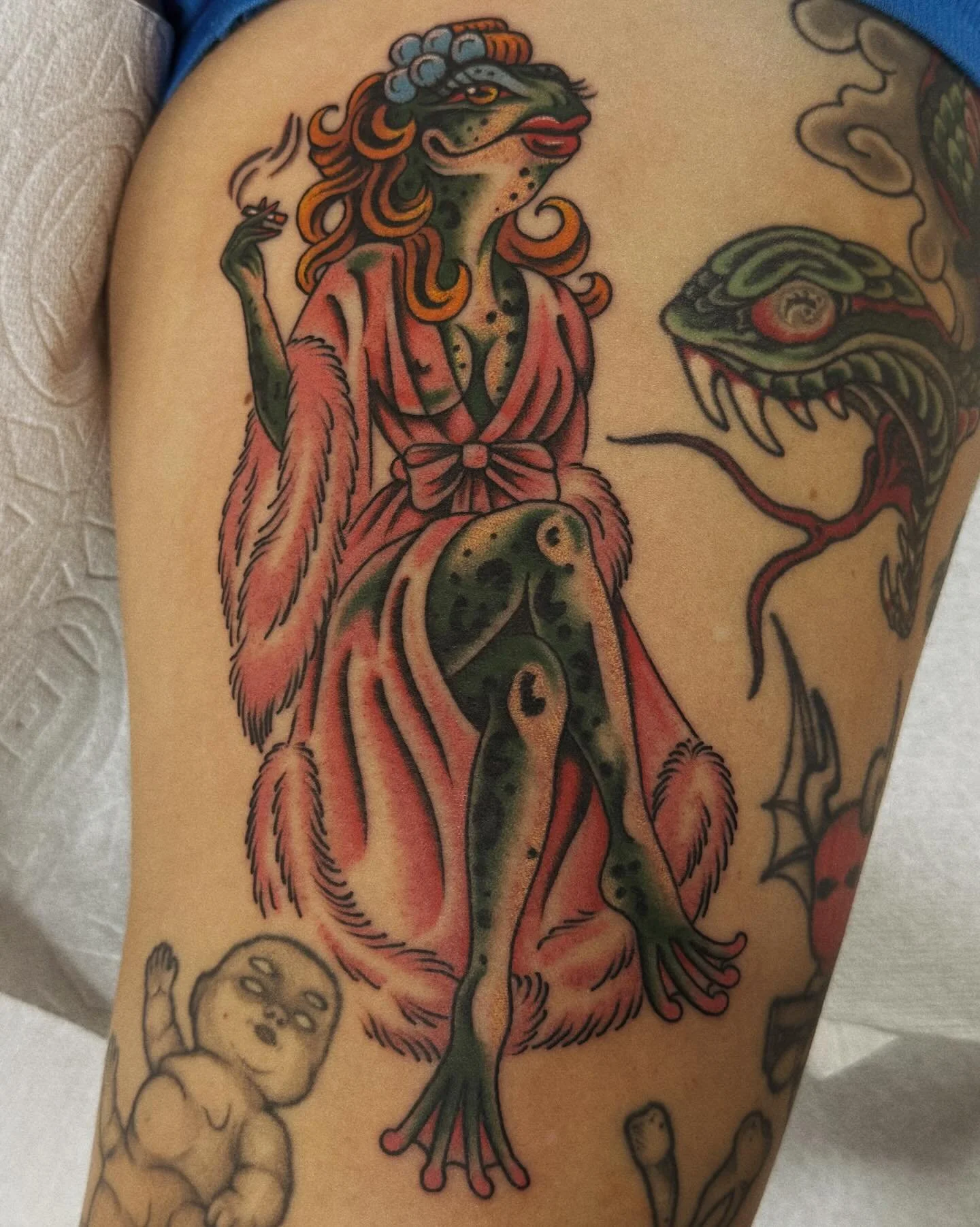 Tattoo of a green anthropomorphic lizard woman with orange hair wearing a pink robe, sitting with one knee up, smoking a cigarette, and surrounded by other tattoo drawings.