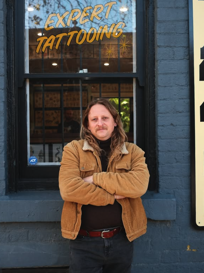 A man with long hair and a mustache standing outside a tattoo shop, crossing his arms, wearing a tan jacket and black pants, with a sign that says 'Expert Tattooing' above the window.