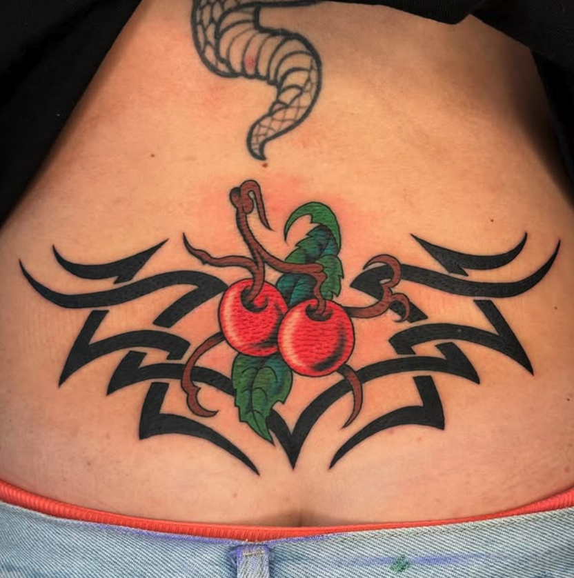 Tattoo of two cherries with green leaves in the center, surrounded by black tribal-style design, with a snake above.