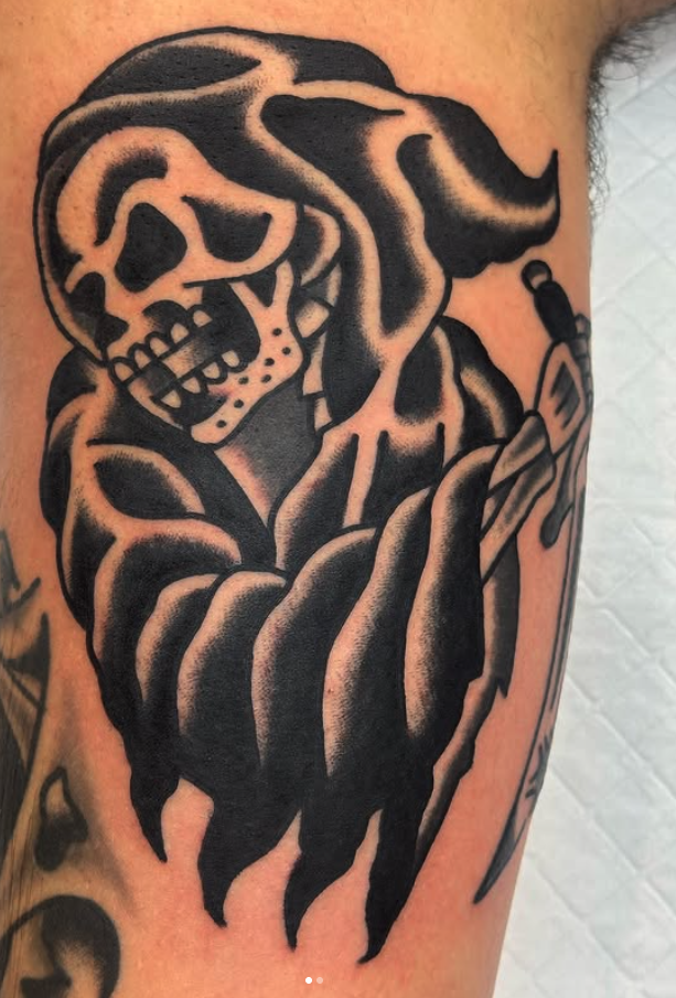 Black tattoo of a skull with a hood covering part of the face, holding a dagger, on a person's arm.