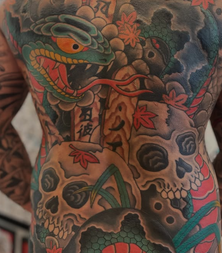 Colorful tattoo of a snake's head and an array of skulls with red maple leaves and Japanese characters.
