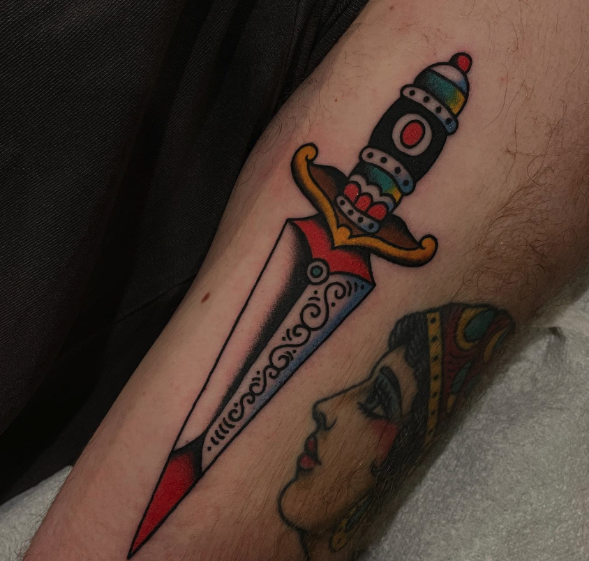 Tattoo of a traditional style dagger with a colorful handle and detailed blade, alongside a side profile of a woman with dark hair and makeup.