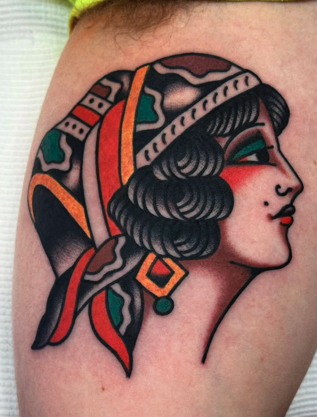 Tattoo of a woman in profile with dark hair, red lipstick, and colorful sheaf on her head, featuring bold lines and vibrant colors.
