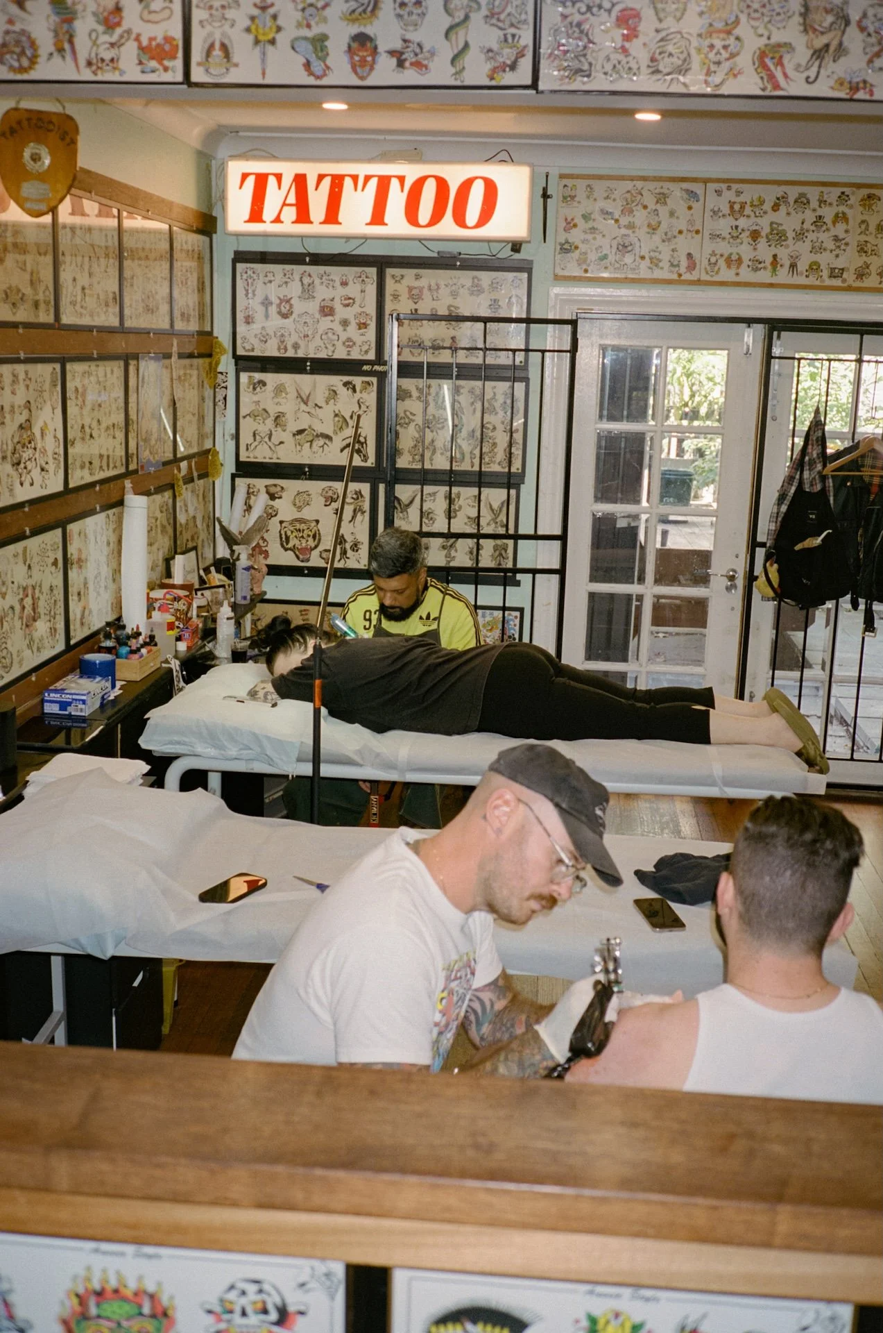 Inside a tattoo parlor with walls decorated with tattoo flash art, showing a tattoo artist working on a client lying on a table. Two other men are seated nearby, one with visible tattoos.