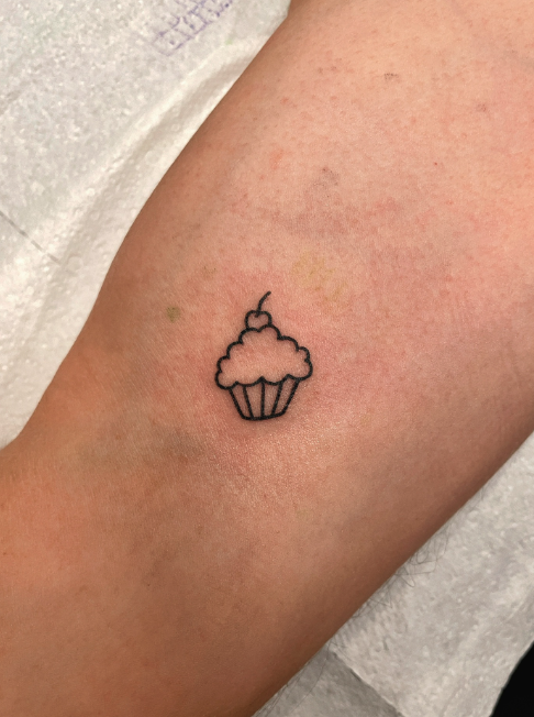 Tattoo of a cupcake with a cherry on top on a person's arm.