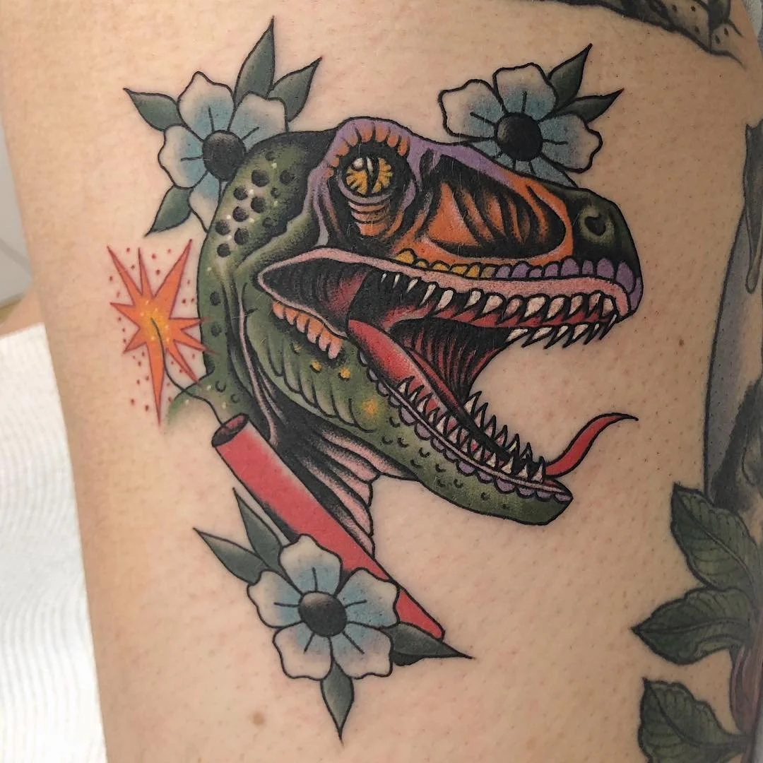 Tattoo of a dinosaur head with an open mouth, sharp teeth, and tongue visible, surrounded by flowers and a small explosion graphic.