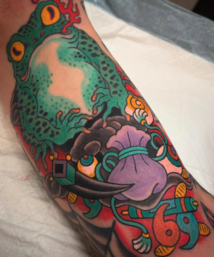 Colorful tattoo of a frog, knife, and abstract elements on an arm.