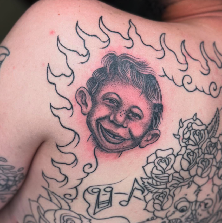 A tattoo of a smiling child's face with freckles, surrounded by smoke-like flames and floral designs.