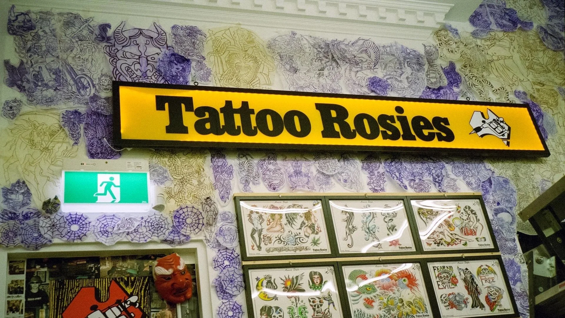 Tattoo shop sign reading 'Tattoo Rosies' with tattoo designs on the wall, surrounded by purple spider web decorations, a green emergency exit sign, and tattoo flash sheets.