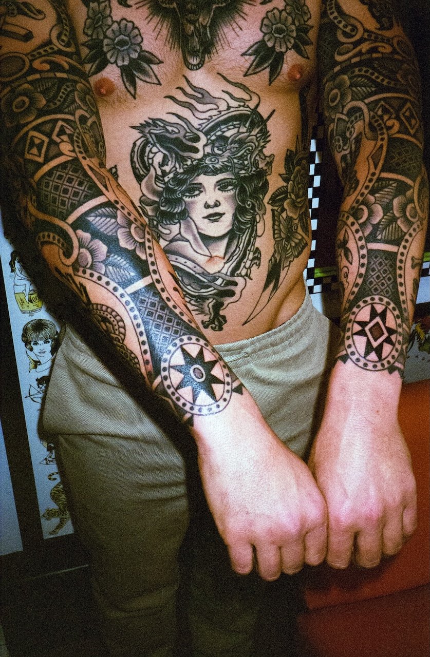 A tattooed man with detailed black ink tattoos on both arms, chest, and stomach, featuring floral patterns and a large portrait of a woman with horned headgear and a dragon. He is wearing beige pants and standing indoors.