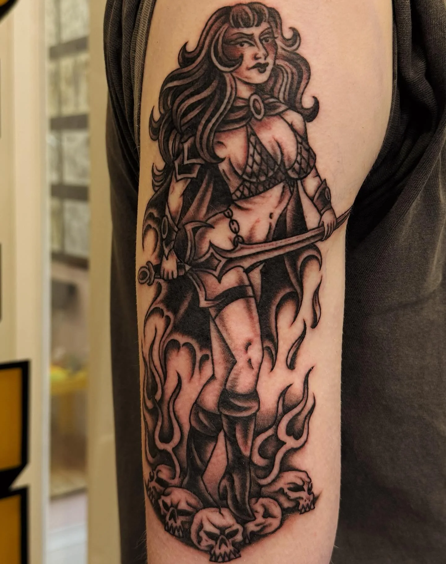 A black and gray tattoo of a fierce warrior woman with long flowing hair, holding a sword, standing on skulls with flames around her.