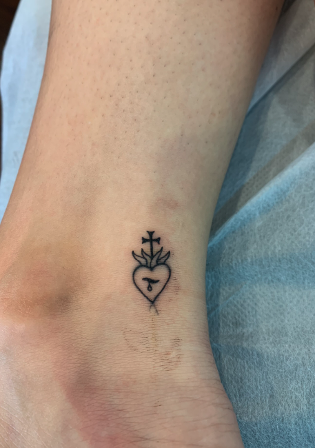 Tattoo of a heart with a small G inside, topped with a flame and a cross, on a person's ankle.