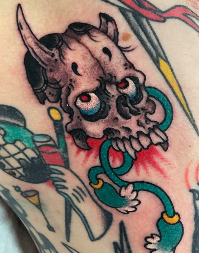 Tattoo of a cartoonish creature with a skull-like face, red eyes, and horns, connected to a tentacle-like appendage with rings.