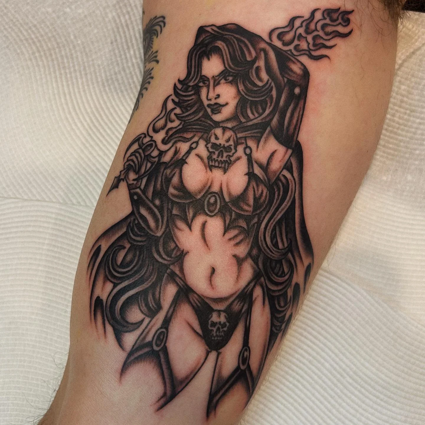 A black and gray tattoo of a woman with long wavy hair, wearing revealing clothing with skull motifs, holding a weapon, and surrounded by flames.