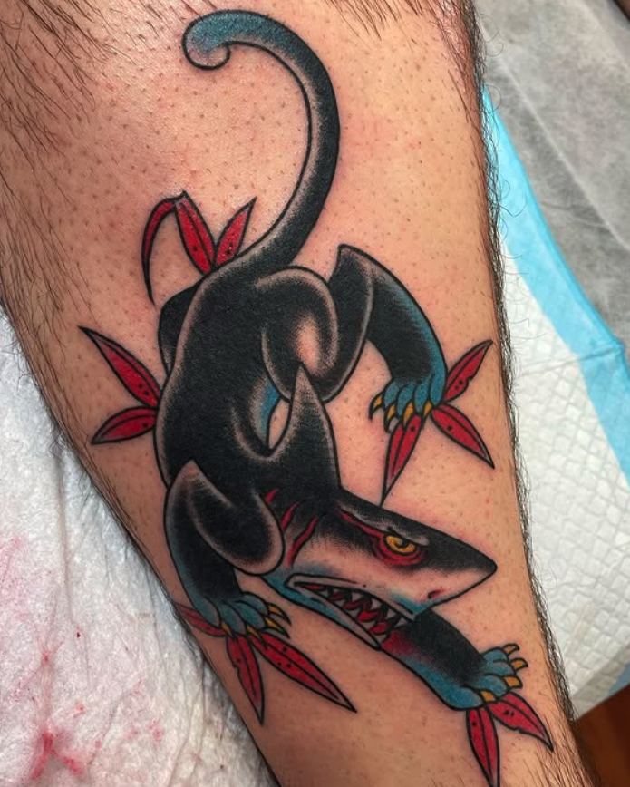 Tattoo of a stylized, aggressive black shark with yellow eyes, red tongue, and sharp teeth, surrounded by red leaves on a person's arm.
