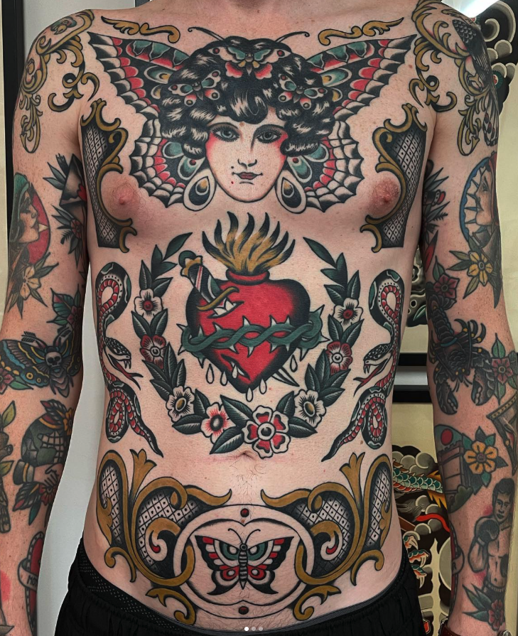 A person's torso covered in colorful tattoos. The most prominent tattoo features a woman's face with butterfly wings and a floral motif surrounding a divided heart with a sword and flames. The tattoos extend to the arms with various floral and animat