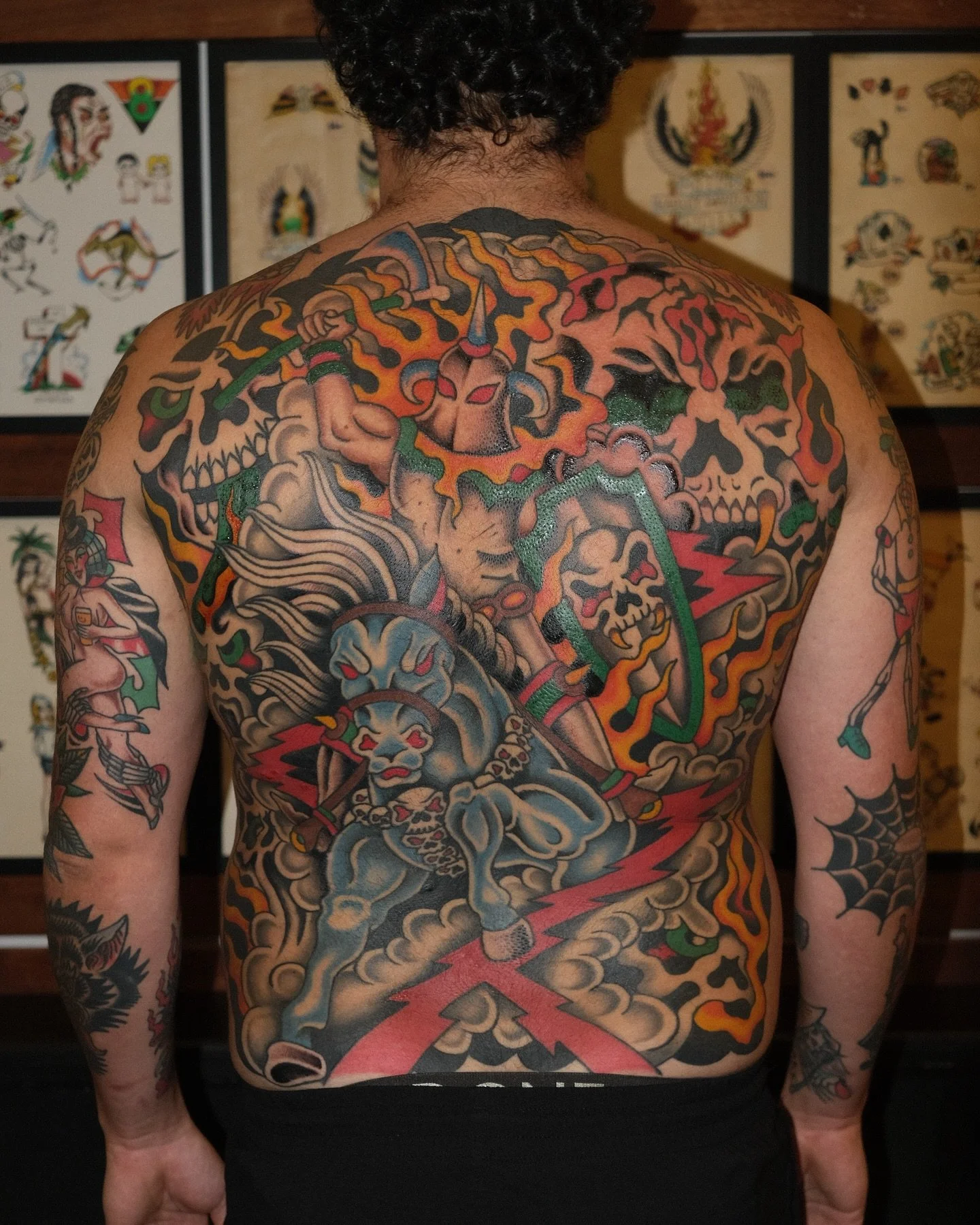 Colorful Japanese-style tattoo covering the person's back and arms, featuring images of a skull, a warrior with a lion mask, flames, clouds, and various traditional motifs.