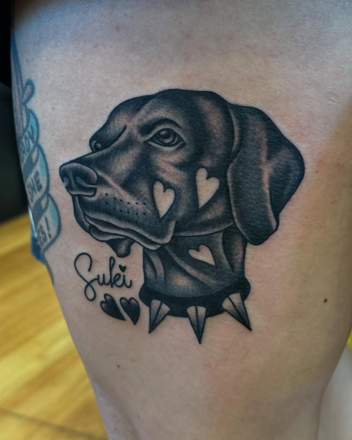 Tattoo of a dog's head with a spiked collar, surrounded by hearts, and the word 'Suki' written below.