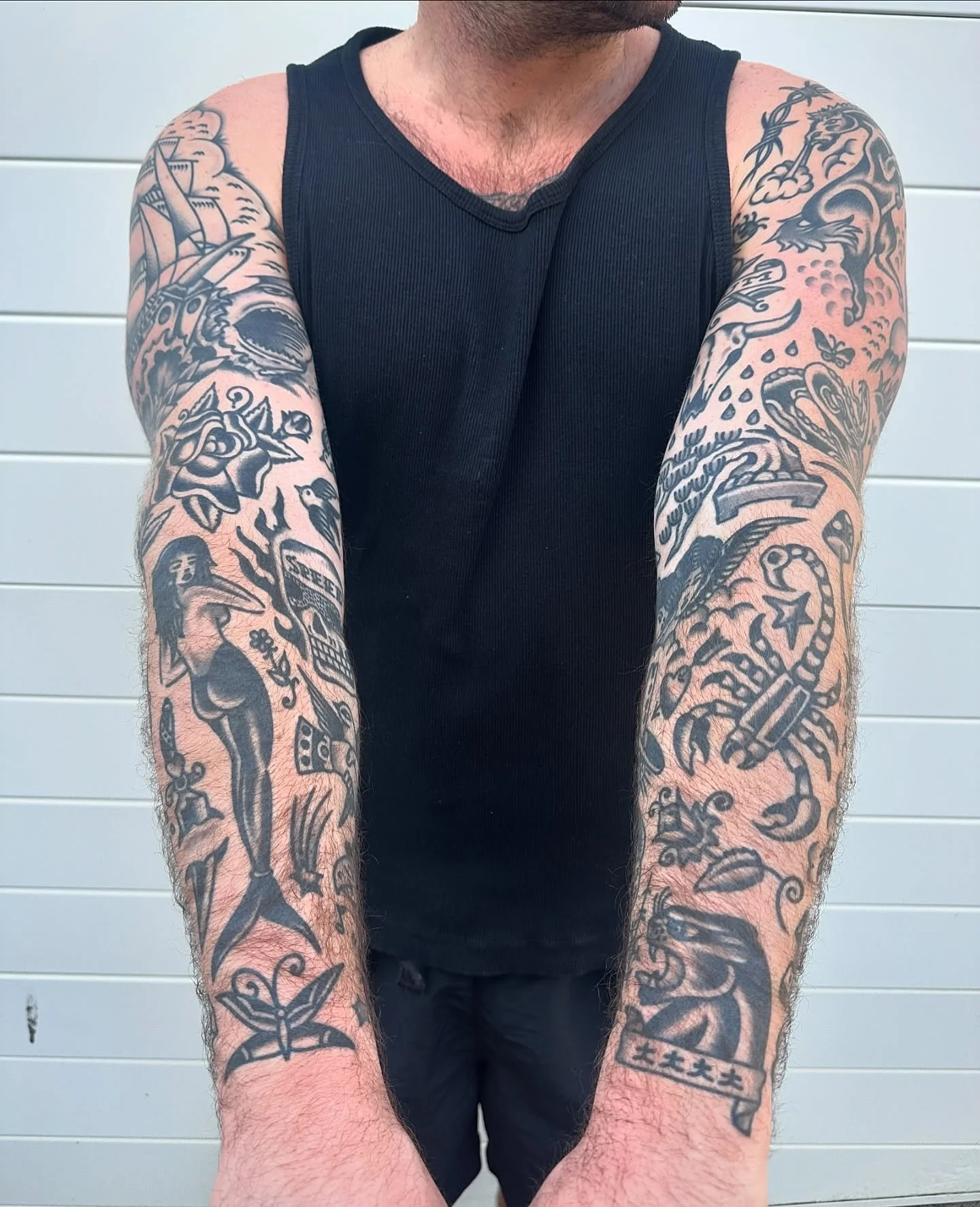 A man with tattoos on both arms is wearing a black sleeveless shirt and standing in front of a white panel background.
