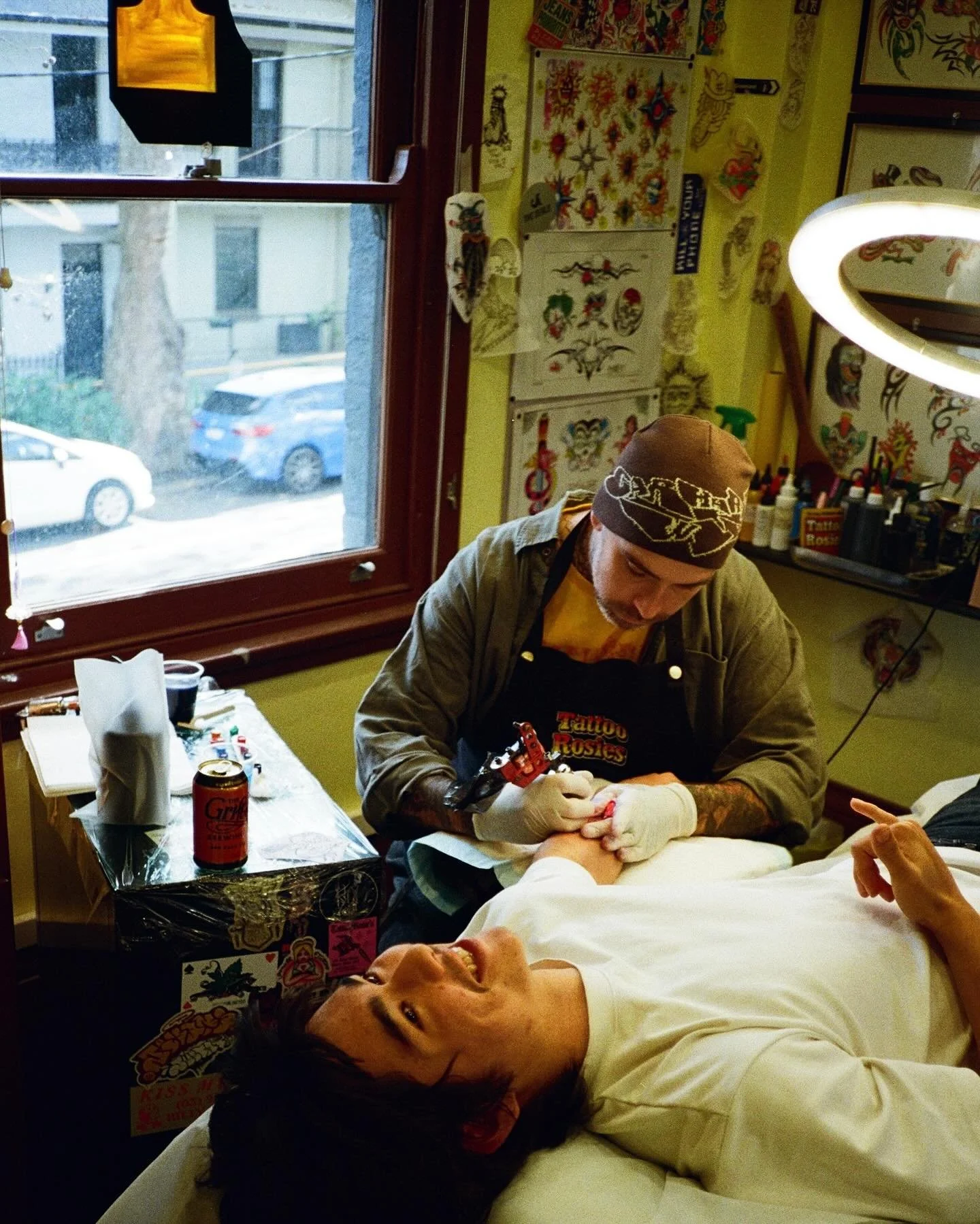 A tattoo artist is working on a client's arm in a tattoo studio, with tattoo designs on the wall behind them and a window showing parked cars outside.