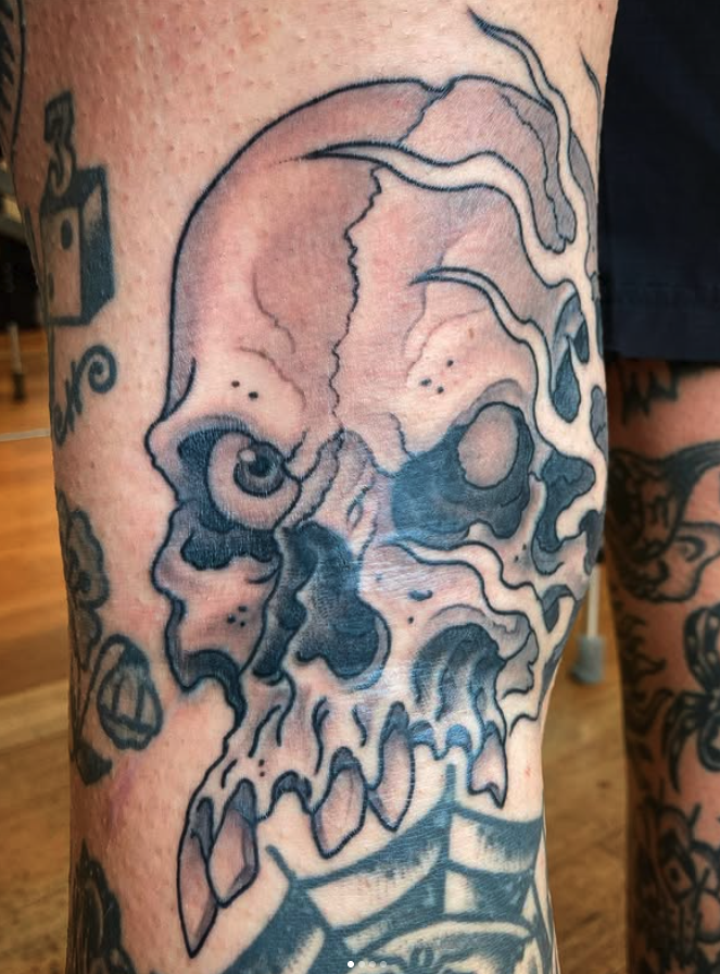 Tattoo of a skull with a distorted, abstract design on a person's thigh, with other tattoos visible on the arm.