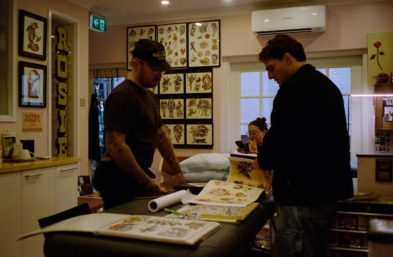 Two men examining tattoo designs on a table in a tattoo parlor, with tattoo flash art on the walls and a woman sitting at a desk in the background, using her phone.