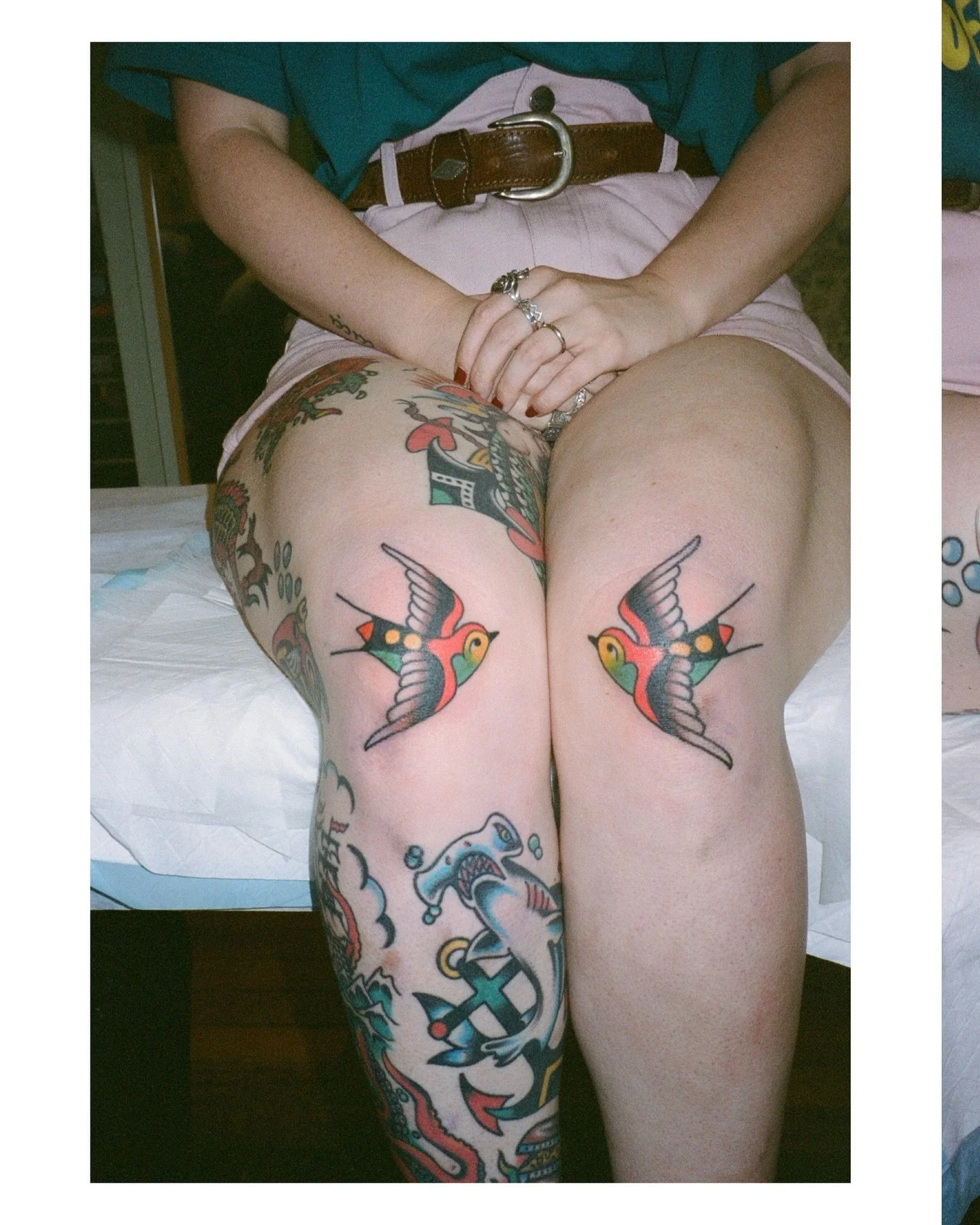 Person showing off colorful sailor-themed tattoos on thighs, with hands resting on lap, wearing rings, bracelets, light pink shorts, teal shirt, and a brown belt.