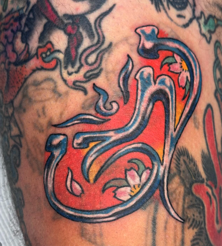 Close-up of a colorful tattoo featuring a stylized, ornate letter 'L' integrated with floral elements, set against a red background on skin.