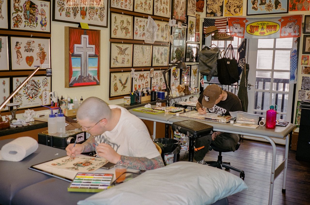 Two tattoo artists working at their stations in a tattoo parlor with walls decorated with colorful tattoo art and framed pictures.