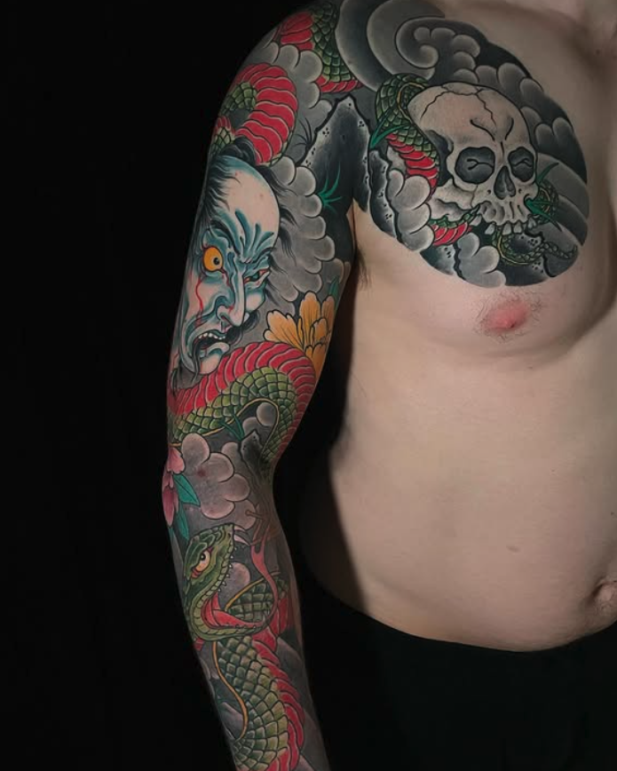 Colorful tattoo sleeve featuring a menacing blue demon face, a green snake, a skull surrounded by clouds, and vibrant flowers on the person's arm and shoulder.