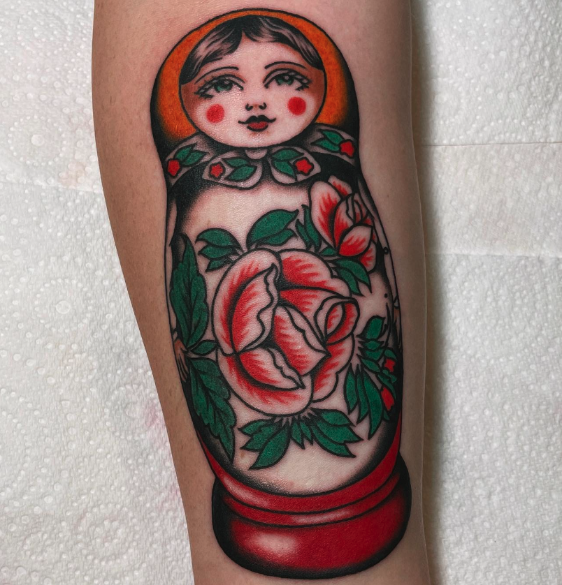 Tattoo of a traditional-style doll with a cute face, rosy cheeks, and a floral design, featuring a large pink and red flower with green leaves, on a person's arm.